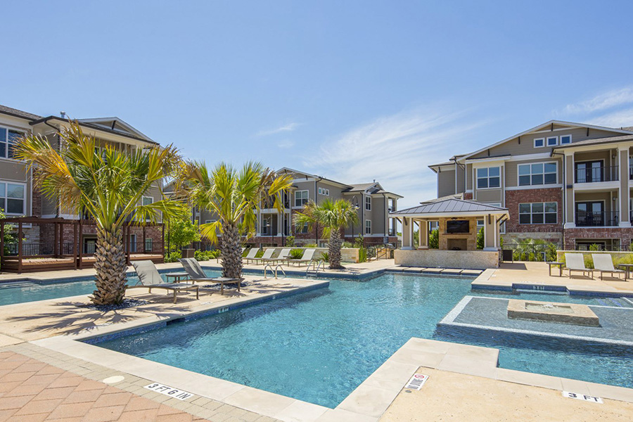 Texas community pool area overlooked by apartments with gazebo including fireplace, TV, palm trees, and lounge chairs.