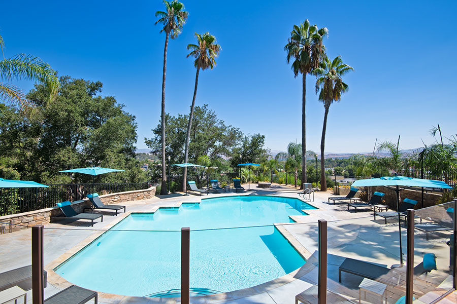 IMT Stevenson Ranch - Raised view of pool area with paver patio, lounge chairs with bright umbrellas, and tall palm trees.