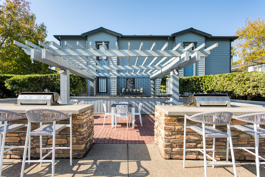outdoor grilling station with brickwork counters with white chairs a white gazebo shades the white patio table and a green bush privacy hedge