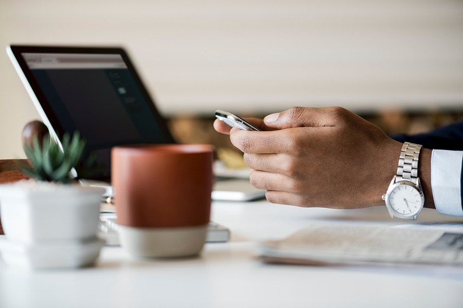 Person with watch holding cell phone at desk with computer, coffee mug, and succulent plants.
