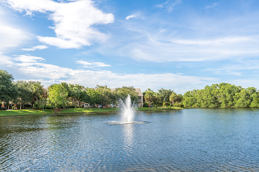 Lake with water fountain lined with green trees along the shoreline under cloudy blue sky.