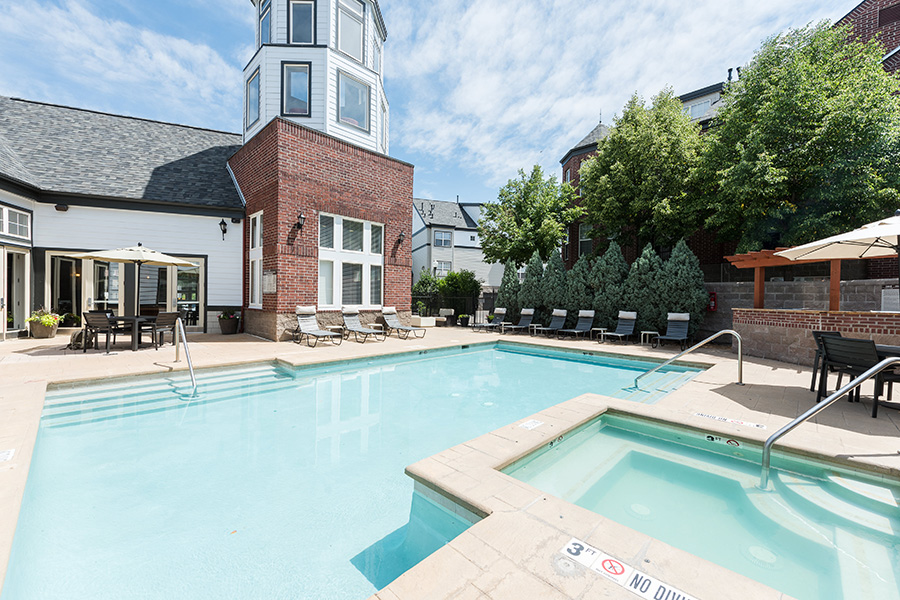 Pool area with clear blue pool and spa, smooth stone pavers, tall trees, and clubhouse with tall tower.