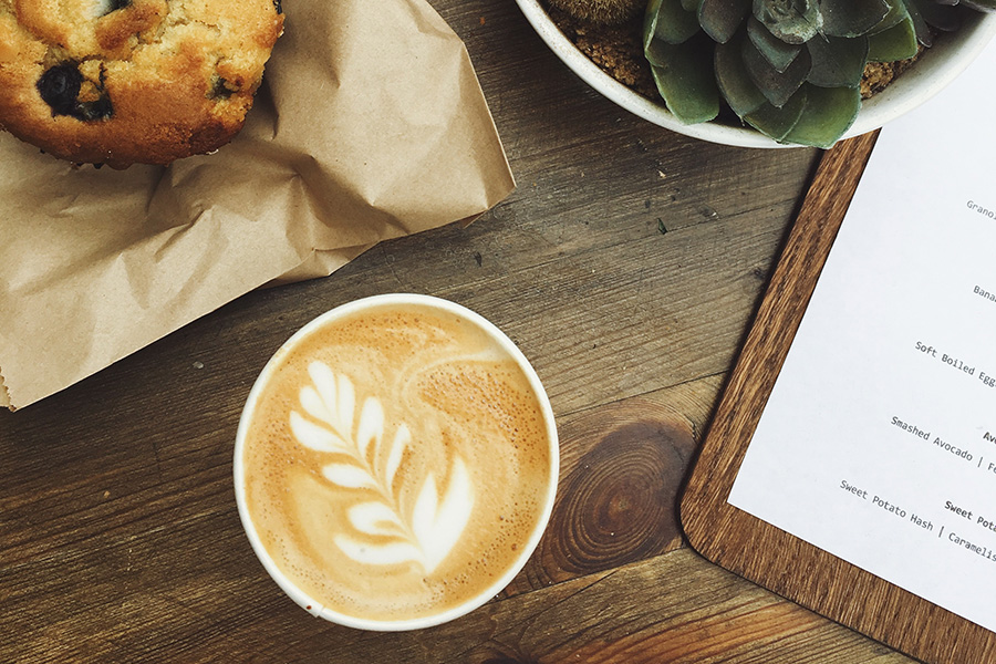 Wood table showing muffin in paper wrapper, potted succulent plant, and cappucino with frothed design.