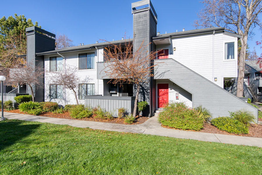 grey and white apartment building with red doors for each unit well kept landscape and bushes around building
