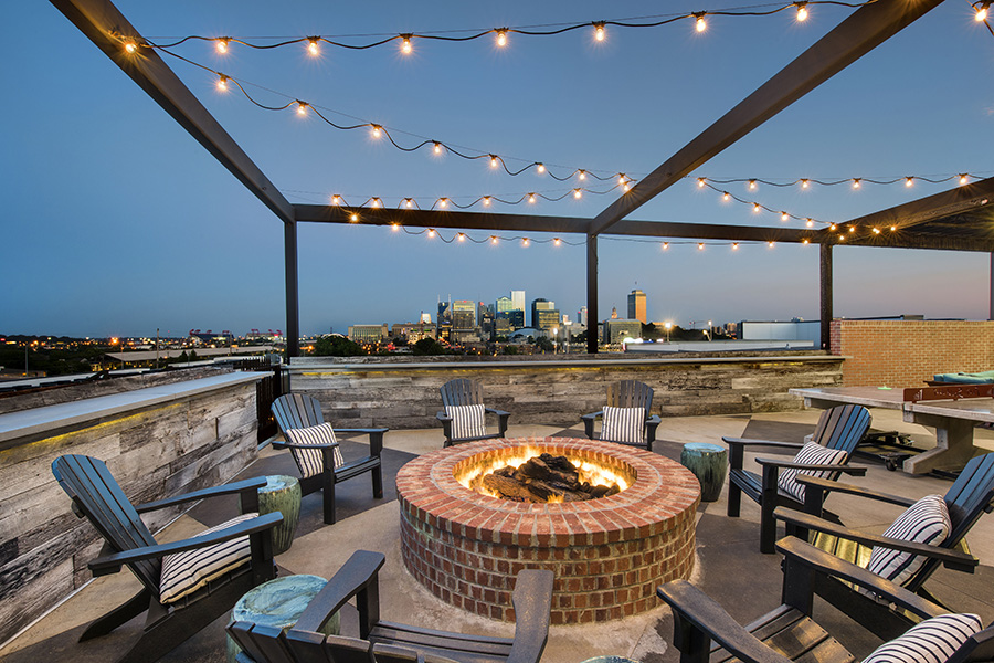 Rooftop lounge with round brick fire pit, lounge chairs, and overhead string lights.
