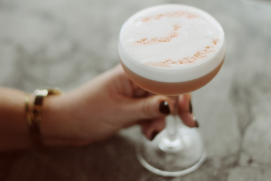 Detail of hand with gold watch holding whiskey sour cocktail on marble bartop.