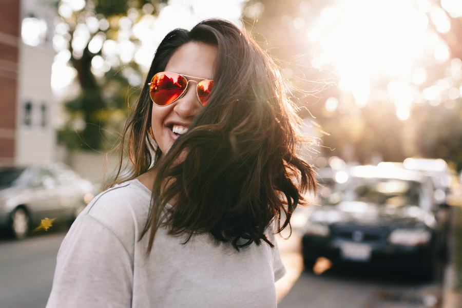 Person with long brown hair and aviator sunglasses stands in street with bright sunlight behind.