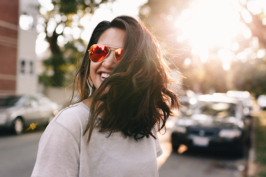 Person with long brown hair and aviator sunglasses stands in street with bright sunlight behind.