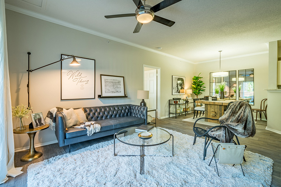Living room with wood floor with rug, wide plush couch, round glass coffee table, and ceiling fan.