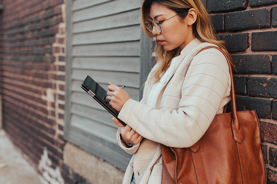 Resident with large leather purse and glasses leans on brick wall and works on tablet with stylus.
