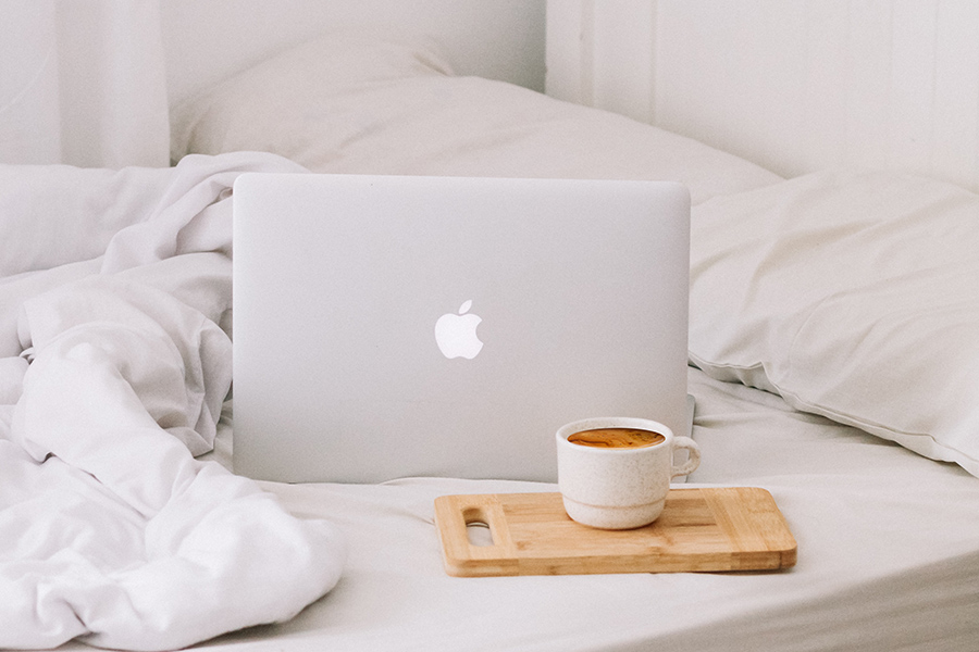 Detail of bed with clean white linens, coffee mug on small wood platter, and silver Mac laptop computer.
