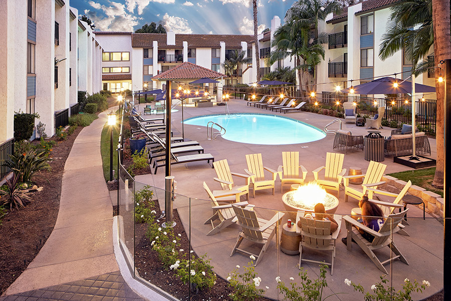 Pool area with fire pit, lounge chairs, umbrellas, and tall palm trees overlooked by apartment balconies.