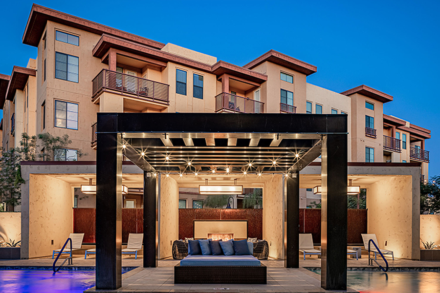 Pool area at dusk with bright lights, daybeds, lounge chairs, and large gazebo.